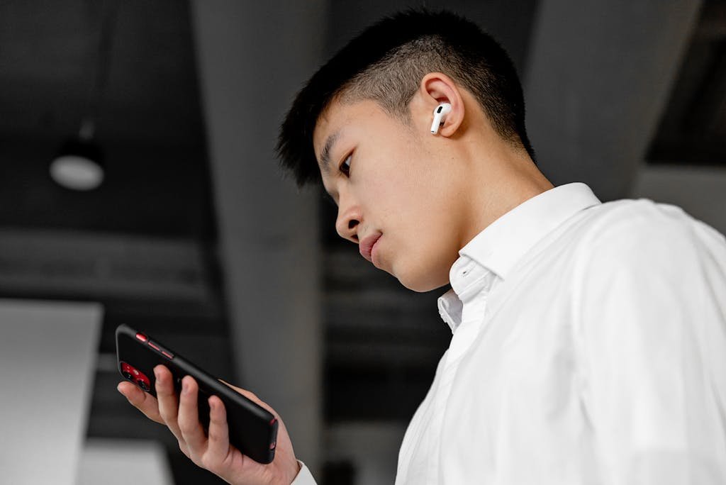 Side view of a young man wearing wireless earbuds and using a smartphone indoors.